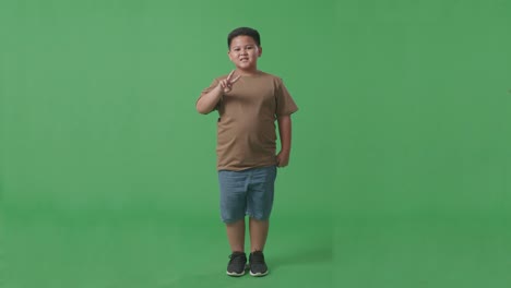 cuerpo lleno de niño asiático mostrando gesto paz y sonriendo mientras estaba de pie en la pantalla verde en el estudio
