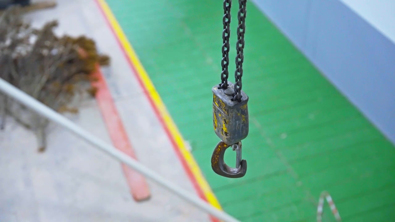 A close-up shot of a weathered metal chain hoist hook suspended from a double chain, with a softly blurred industrial background in green, blue, and orange tones.