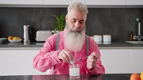 A-serious-elderly-man-with-gray-hair-and-a-lush-beard-in-a-pink-shirt-pours-white-medicine-into-a-glass-and-stirs-it-with-water-using-a-spoon-in-a-modern-kitchen