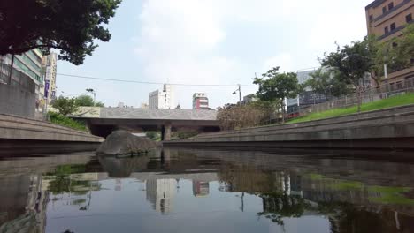 Quiet-urban-river-with-reflections-under-a-bridge,-greenery-around,-city-buildings-in-the-background,-cloudy-day