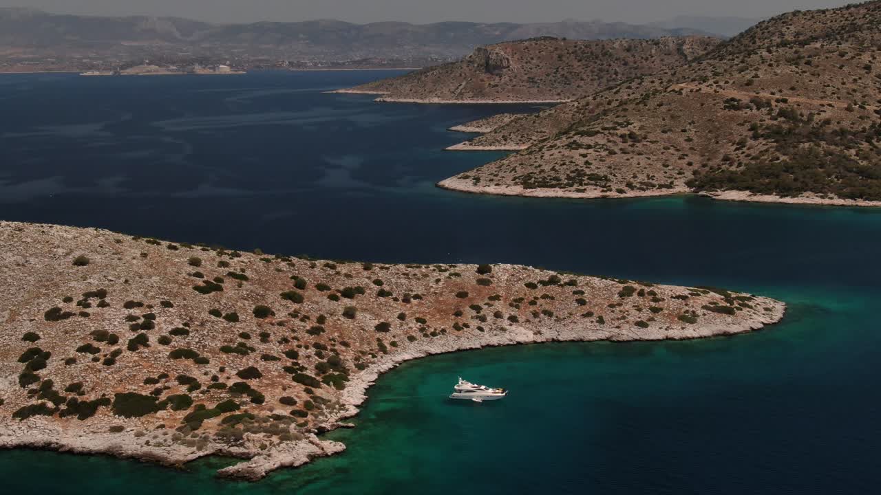 Drone video captures a serene aerial view of a boat anchored near the rocky coastline of Amorgos Island, Greece. The gradient turquoise waters contrast against rugged cliffs in stunning beauty