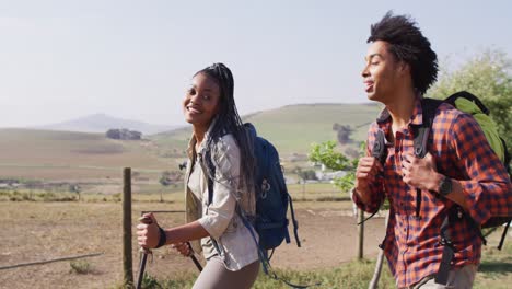 Happy-african-american-couple-with-backpacks,-hiking-with-trekking-poles-together,-slow-motion