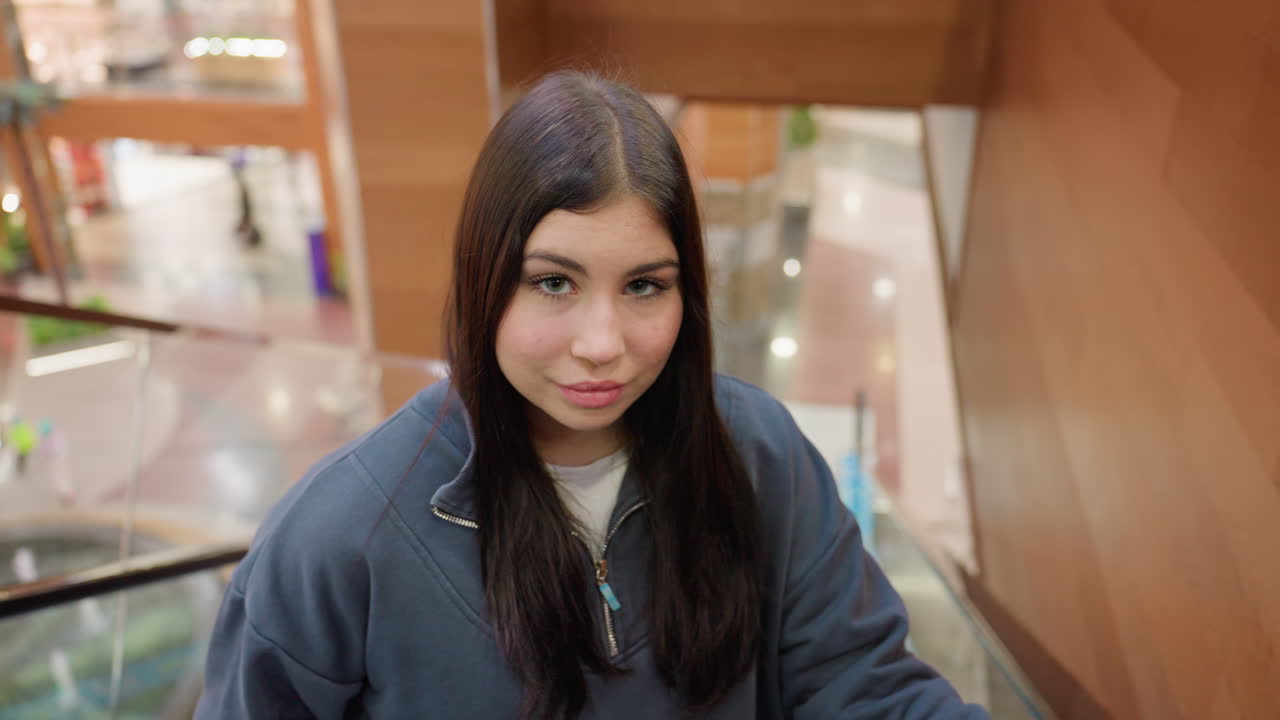 Young woman with long dark hair descending escalator inside shopping mall, looking directly at camera with soft elegant expression, modern background