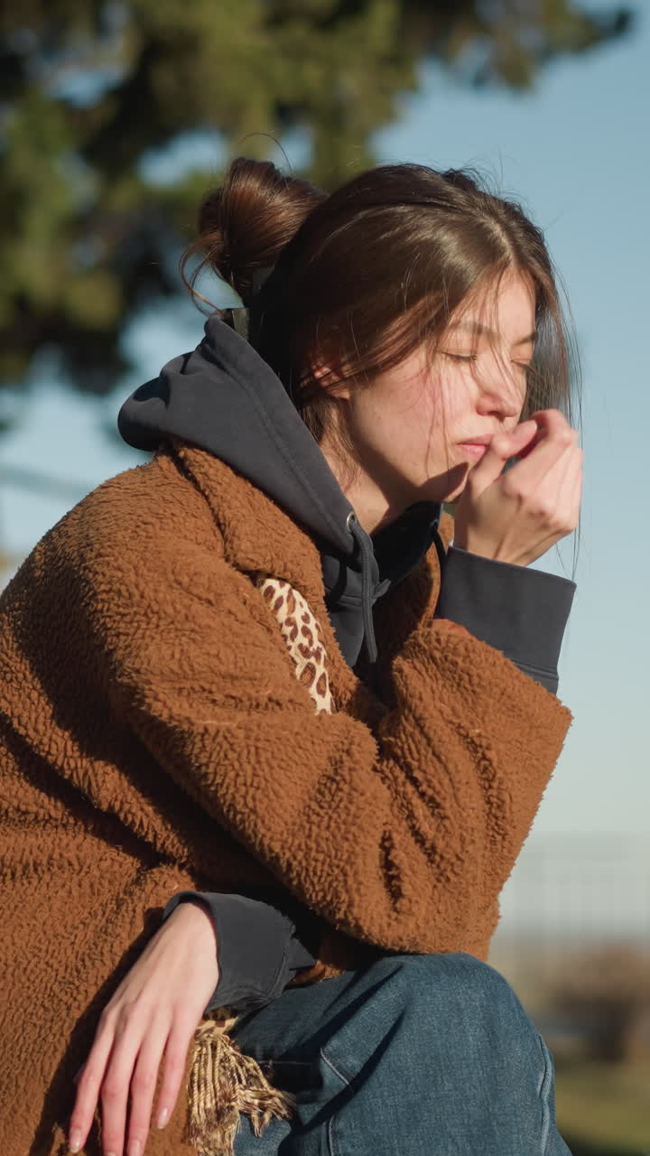 한 소녀가 도시 공원의 벤치에 혼자 앉아 슬에 싸여 그녀의 얼굴에 손을 고 있습니다. 그녀는 갈색 코트와 파란 청바지를 입고, 그녀의 에 검은 배을 배치합니다.