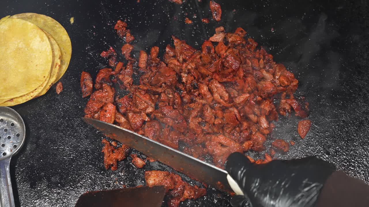 Riprese dall'alto di carne al pastor che sfrigola su una griglia rovente mentre un venditore taglia e sminuzza i pezzi con un coltello e una spatola. Vapore e rosolatura trasmettono calore, aroma e la freschezza della cucina di strada