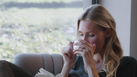 woman with hot drink relaxing in chair by window at home reading book