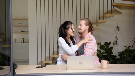 a biracial young woman and caucasian young man sit with laptop in kitchen at home