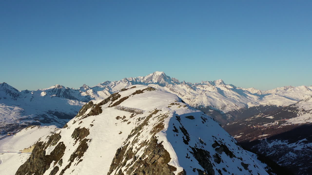 Ripresa con drone che si muove da destra a sinistra, mostrando una cima montuosa innevata con il Monte Bianco sullo sfondo, in una giornata di sole invernale.