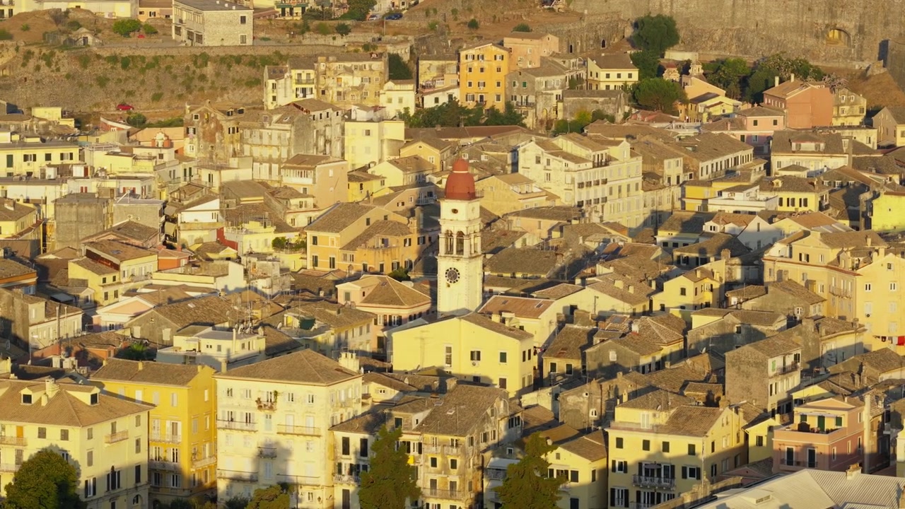 Lenta panoramica all'ora d'oro sulla città vecchia di Corfù con edifici densi e il campanile della chiesa di San Spiridione nella calda luce della sera.