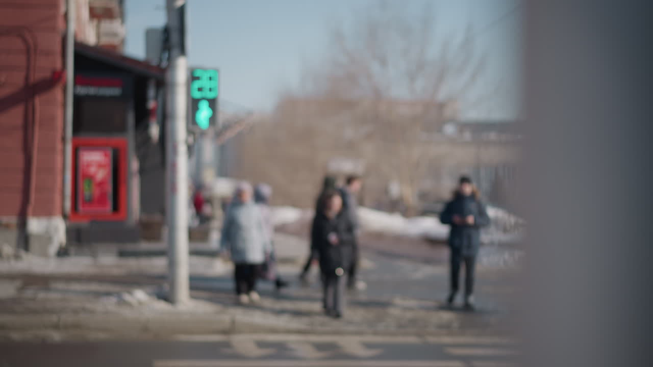 blur view shows pedestrians waiting then crossing city street at signal, red icon glowing near corner storefront, winter snow along curb, soft bokeh background, urban commute moment with motion
