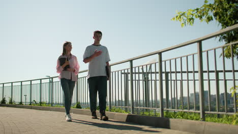 niña con camisa rosa sosteniendo un cuaderno caminando junto a un amigo masculino en un camino pavimentado cerca de una barandilla de hierro, participando en una conversación alegre rodeada de vegetación, paisaje urbano y arquitectura urbana