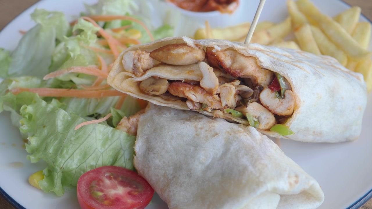 Hand Lifting a Chicken Wrap from a Plate with Salad and Fries