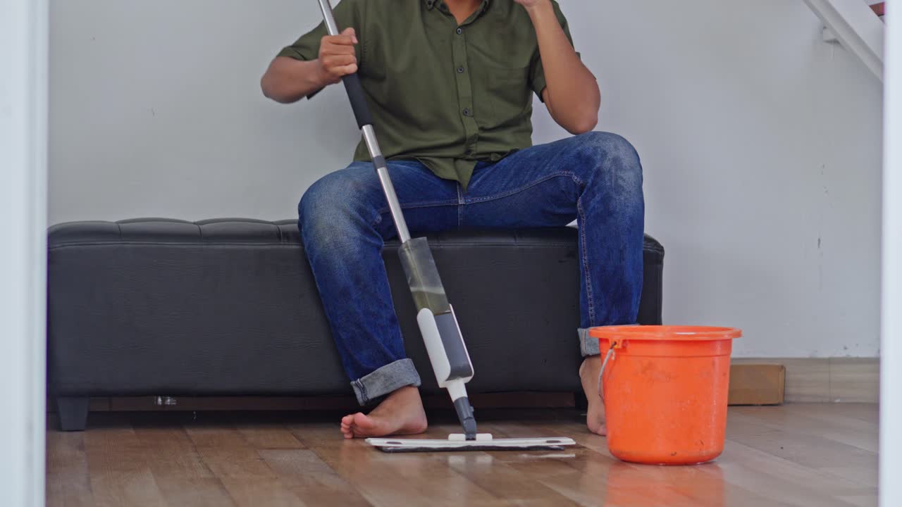 Ung mann sitter på benk og ser frustrert ut mens han mopper ved siden av en oransje bøtte på tregulv