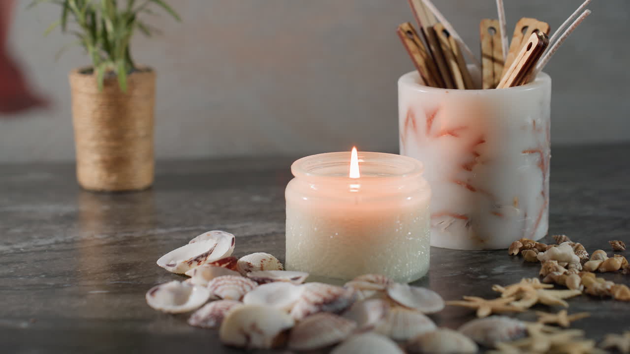 White candle burning steadily in glass jar surrounded by seashells starfish and small snail shells on dark marble surface soft glow creating peaceful cozy serene elegant atmosphere