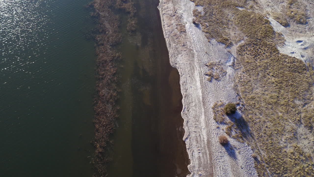 Top-down drone fly over a highland riverbank shoreline with sand and lake water meeting under sunlight in Patagonia