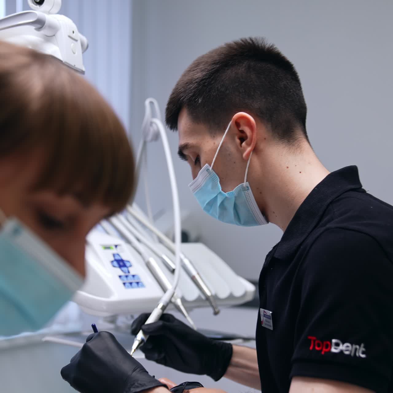 Giovane dentista moro in mascherina che lavora nel suo studio. Il dottore sistema la posizione della lampada e prende il trapano. Infermiera in primo piano