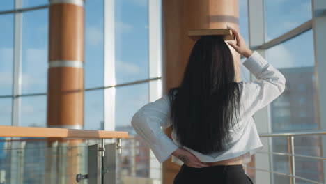 back view of young girl in white shirt walking with book on head while turning and scratching waist, long dark hair cascading down, inside sunlit modern space with large windows and warm architecture