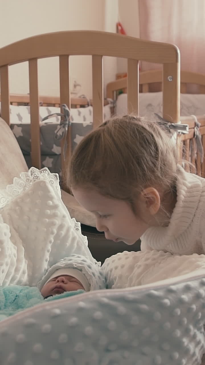 funny little girl in sweater looks at sleeping junior newborn brother in comfortable swaddle envelope on sofa