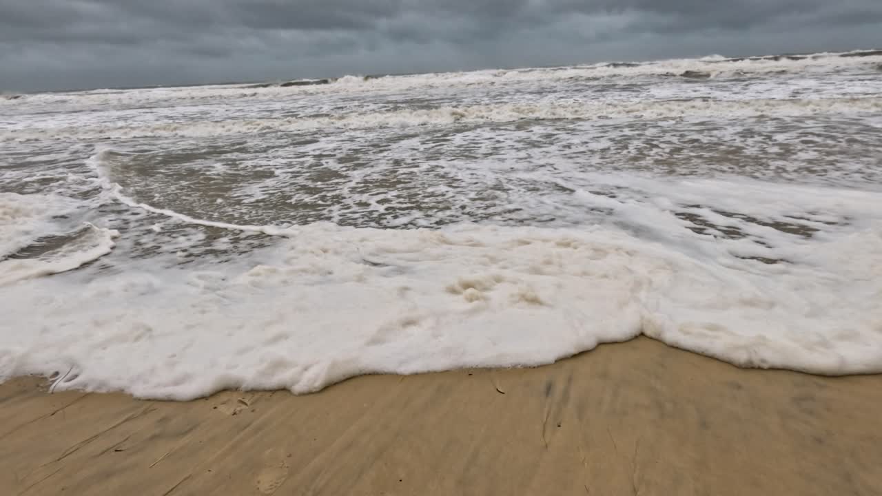강력한 바다 파도가 모래 해변에 부히며 구름이 가득한 하늘 아래에서 거품 모양의 패턴을 만들어 자연의 원시 에너지를 포착합니다.