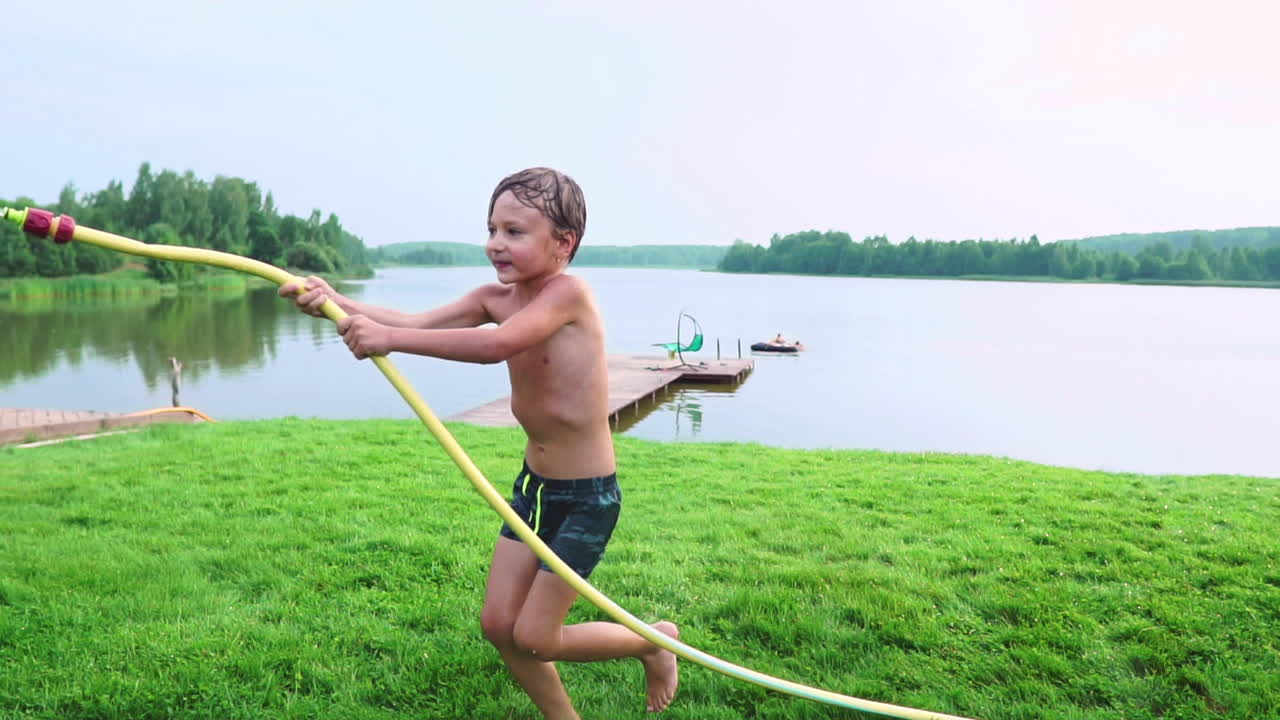 여름에 수영복을 입은 소년은 동생에게 물을 부어 호수 근처의 잔디 ⁇ 에서 공원에서 재미를 느끼고 있습니다.