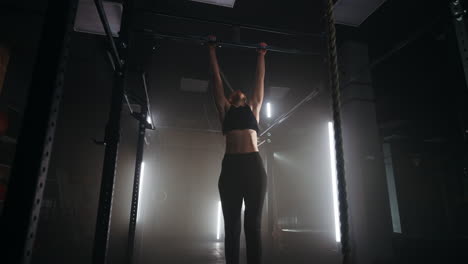 athletic woman is doing pull-ups on crossbar in gym training alone in dark fitness hall in night