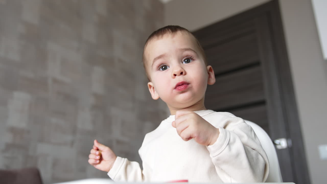 Lovely Caucasian kid sitting at the feeding table. Baby boy taking the pieces of food from plate and putting them into mouth. Low angle view close up.