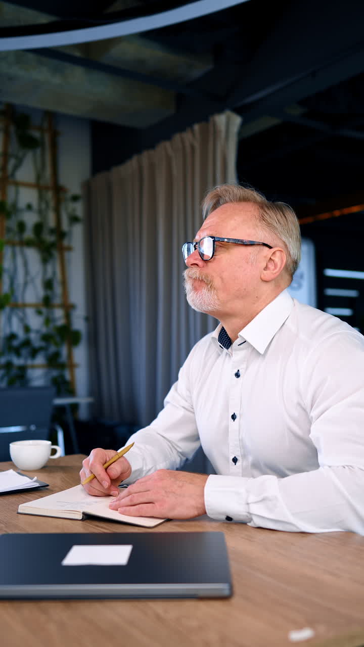 Pensive Caucasian mid-aged man sits at desk in the office. Businessman creating ideas, checking information on laptop. Vertical video.