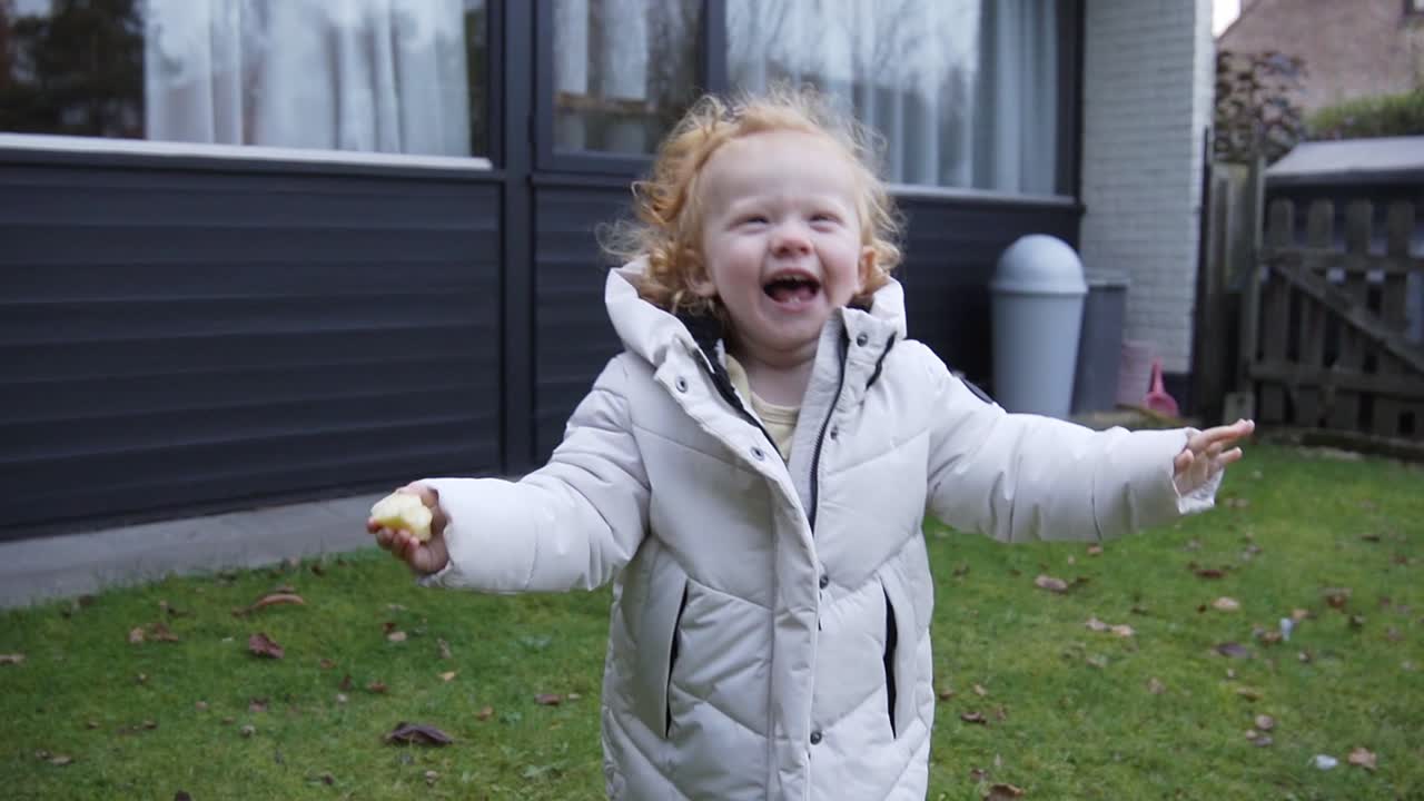 Juokseva taapero talvitakissa isolla hymyllä puutarhan läpi