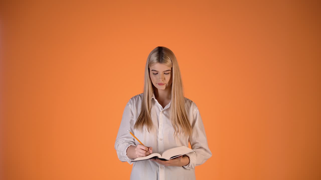 Fokusert ung blond kvinne, som skriver notater i en papirnotatbok, fargerikt studiobilde.
