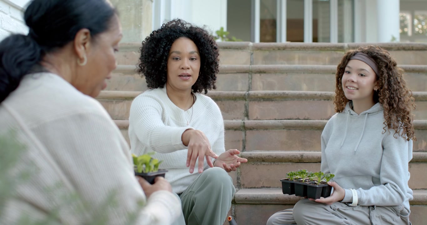 African American female family on stoop, adult woman handing seedling tray, using trowel, teaching
