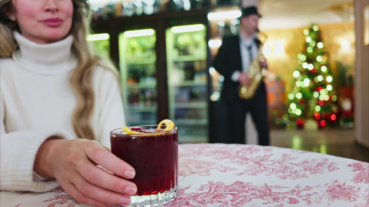 Close up of a woman holding a negroni cocktail on a red and white tablecloth at a restaurant with a Christmas tree on the background