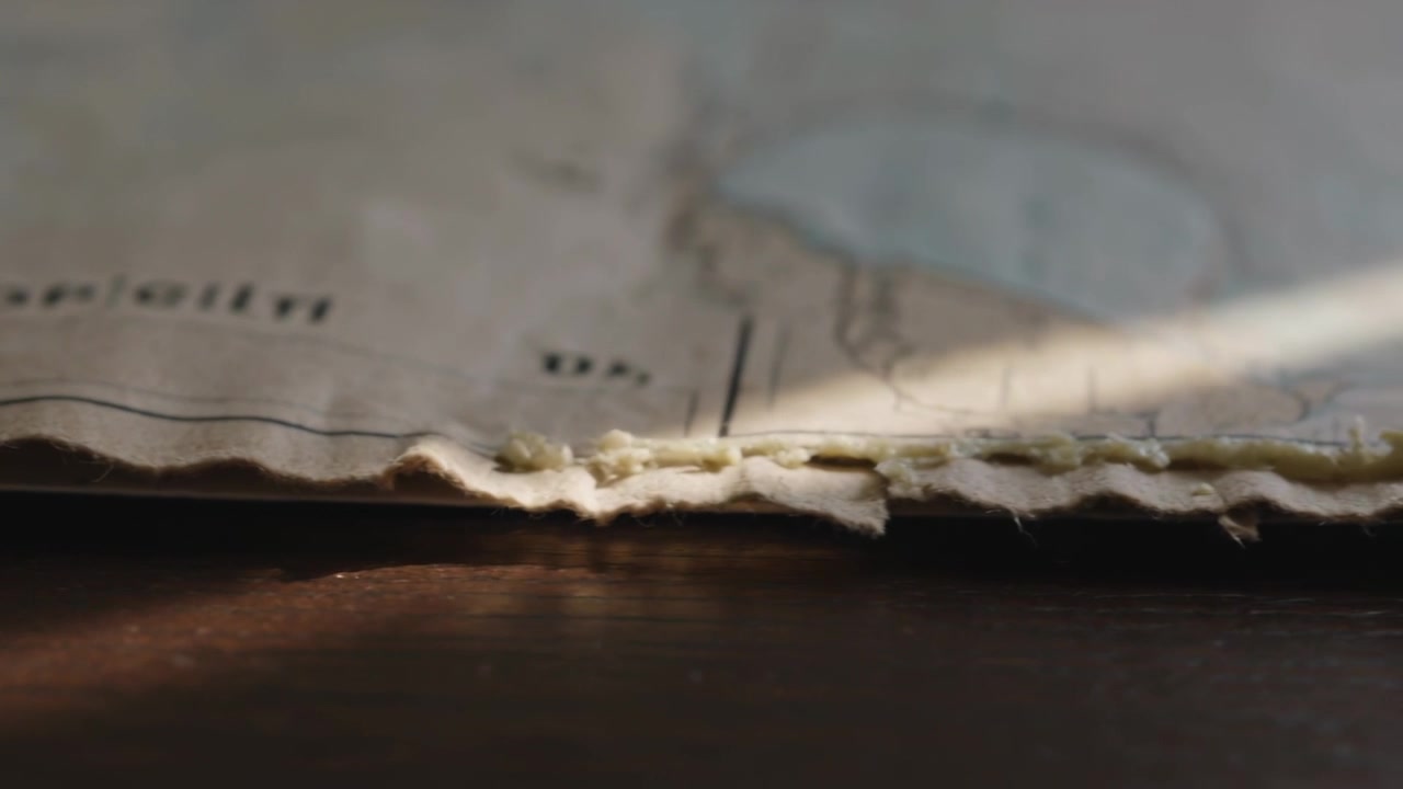 Shifting slanted sunbeam crossing torn stacked map on dark wooden tabletop, revealing stitch line
