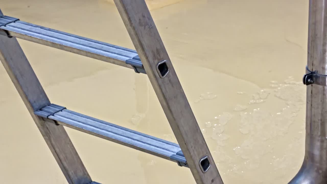 Plano tilt-up de uma escada de alumínio com pés de borracha em piso industrial revestido com epóxi amarelo, ao lado de uma tampa de bueiro aberta e equipamento de aço inoxidável com poças e gotas de água
