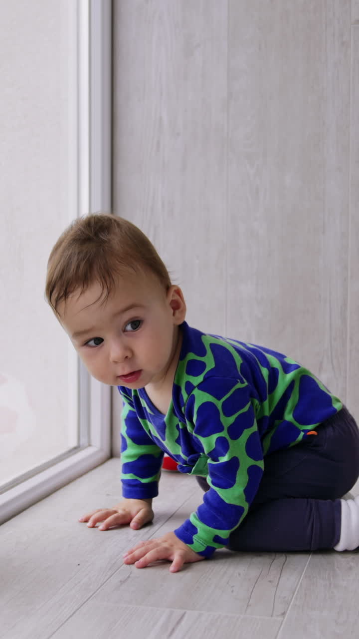 Lovely Caucasian baby looks curiously into window. Beautiful little boy moving by the floor in the room. Vertical video