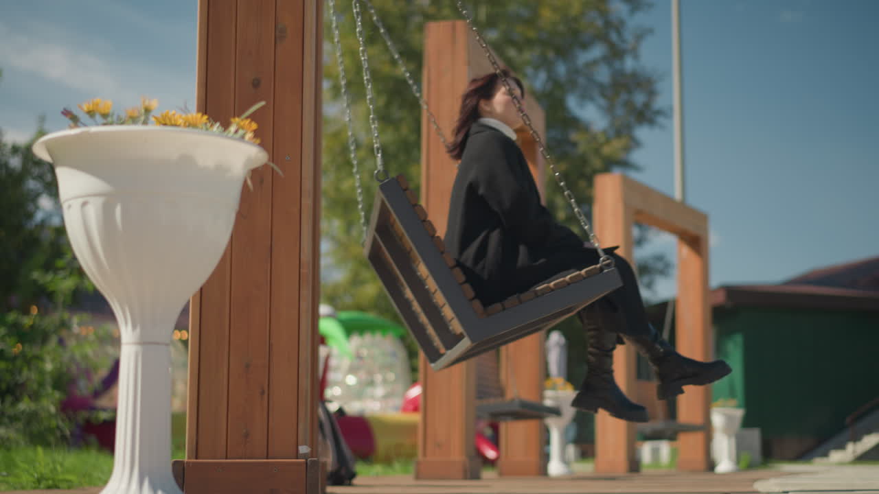 화려한 하늘 아래, 빛이 빛나는 공원, 초록색과 장식적인 꽃으로 둘러싸인, 멋진 검은 코트와 부츠를 입은 여자가 나무 스에서 휴식을 취하고 있습니다.