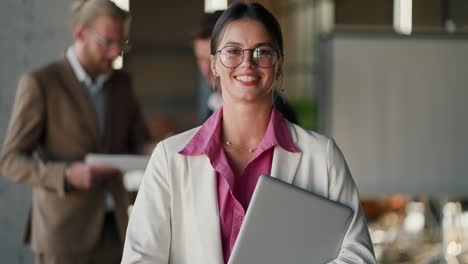 retrato de una chica morena con gafas redondas en una chaqueta blanca y camisa rosa que está de pie con una computadora portátil y ajusta sus gafas contra una oficina moderna y sus colegas