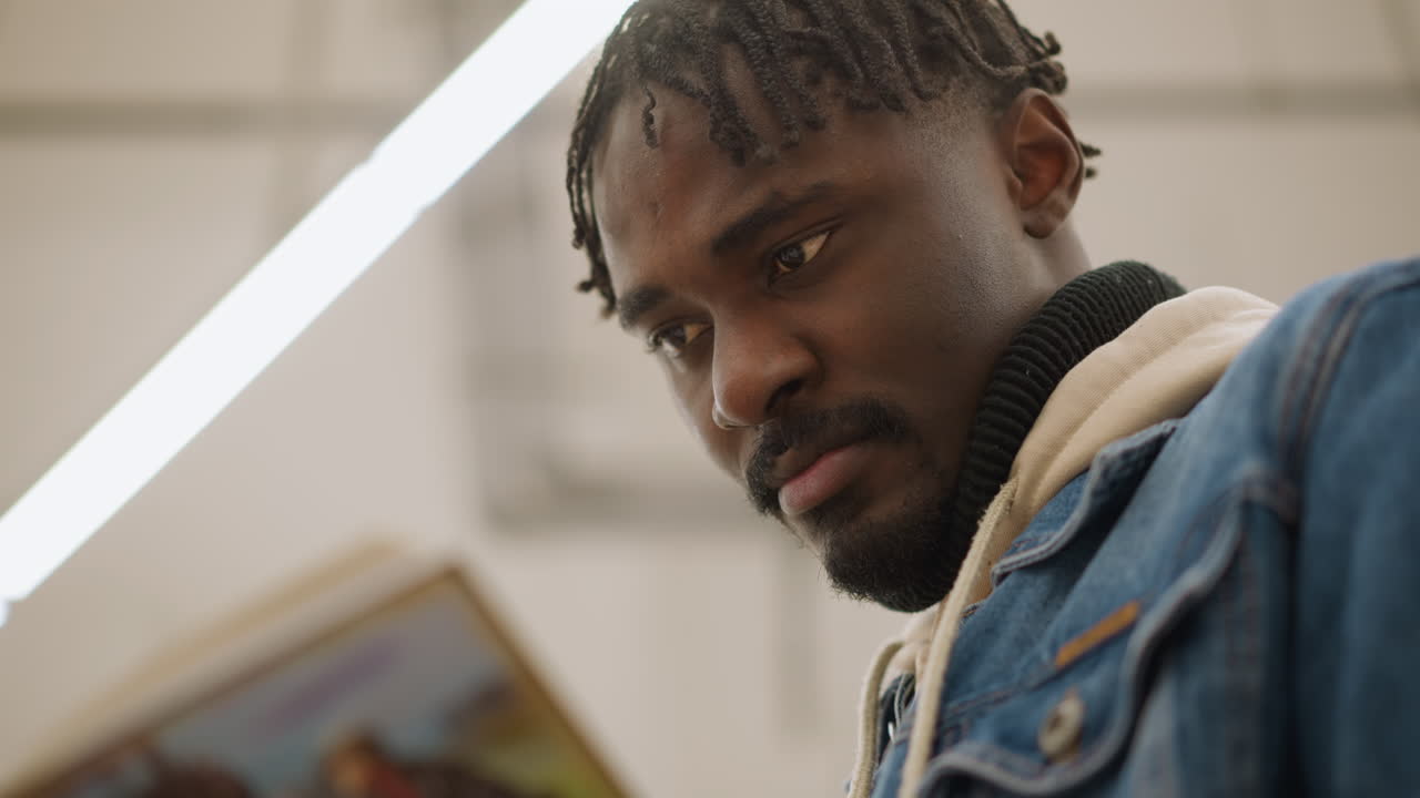 Male in denim jacket focused on reading book closely in modern library. He examines pages with concentration, surrounded by well-organized bookshelves and colorful books in peaceful library setting