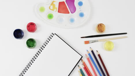 vista aérea del cuaderno de notas, pinturas, pinceles y lápices de colores sobre fondo blanco, en cámara lenta
