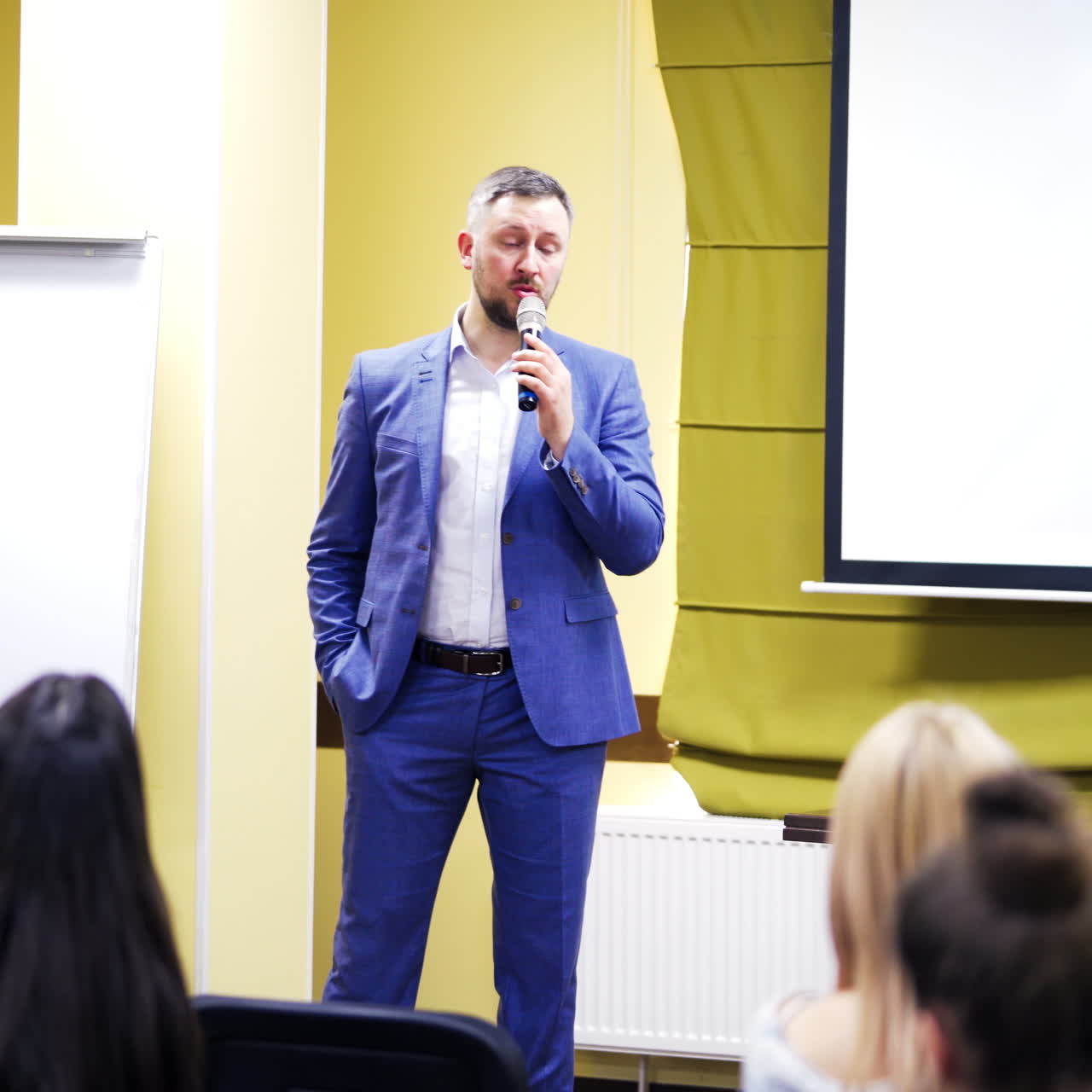 Speaker in blue suit talking to a microphone at conference. Handsome businessman shares his experience with audience indoors. Professional ideas