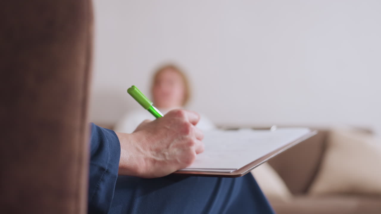 Practitioner takes notes with green pen on clipboard during therapy session, while client is seen blurred in background engaged in conversation, illustrating active listening in calm environment