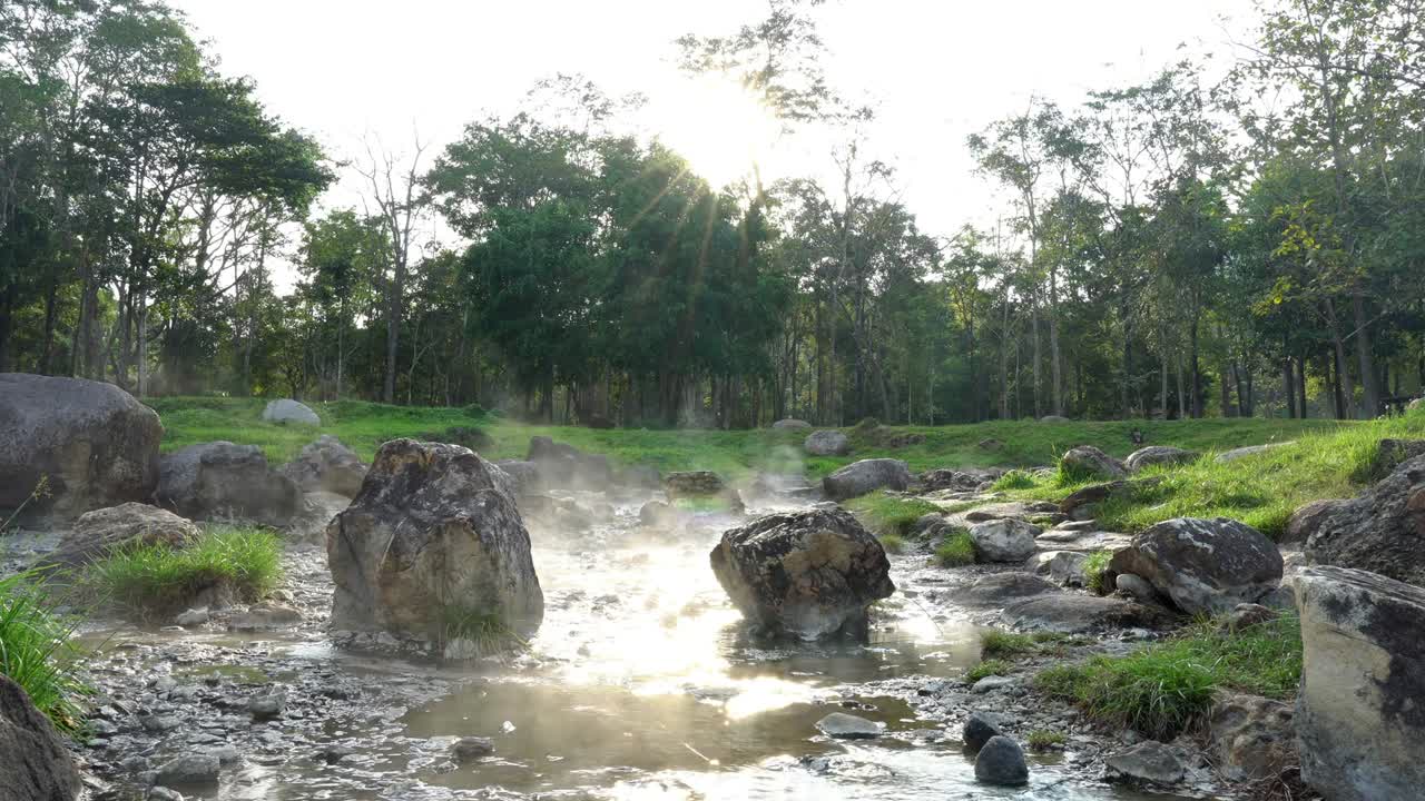 zoom lento de 4k en la toma de aguas termales naturales que fluyen a lo largo de las rocas bajo el reflejo de la luz solar y las llamaradas solares, el vapor flotando con árboles y hierba de fondo. lampang, tailandia.