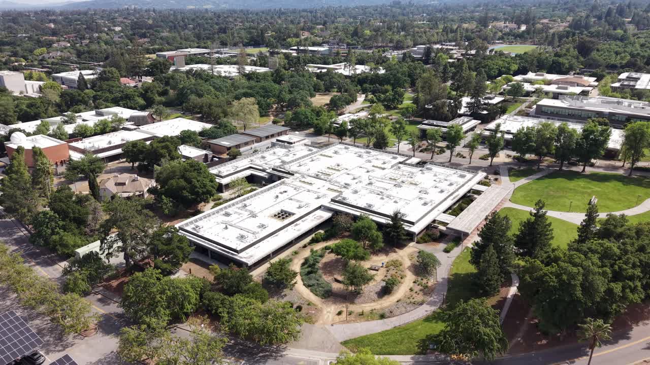 Aerial drone footage zooming out of a modern building on the West Valley College campus, highlighting its architecture and the surrounding green landscape of Saratoga, California on a sunny day