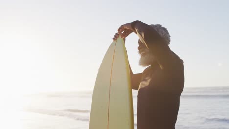 Happy-senior-african-american-man-holding-surfboard-on-sunny-beach