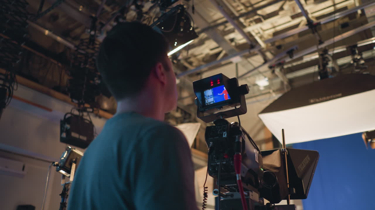 Cameraman operating professional video camera during studio recording session with presenter in red dress on blue screen background under ceiling rig lights and soft boxes in high tech production setup