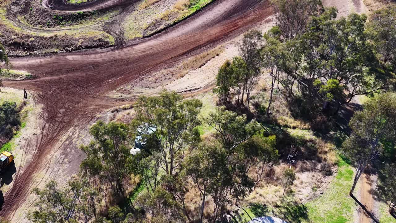 Drone glides from paved road over trees to dirt motocross track in bright daylight