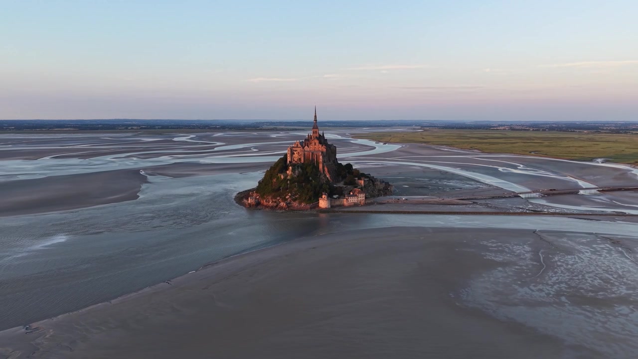 Wide cinematic drone orbit around Mont Saint-Michel Abbey at sunset, showcasing the tidal island surrounded by expansive mudflats and winding waterways in Normandy, France