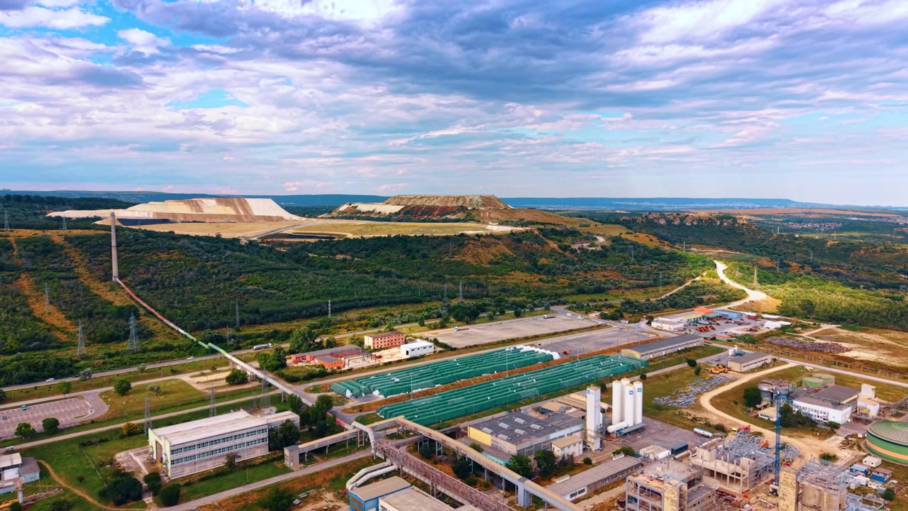 Processing plant below terraced quarry mounds and layered clouds. Aerial panorama includes conveyors, tanks, pipelines, workshops and vegetation across a broad valley.