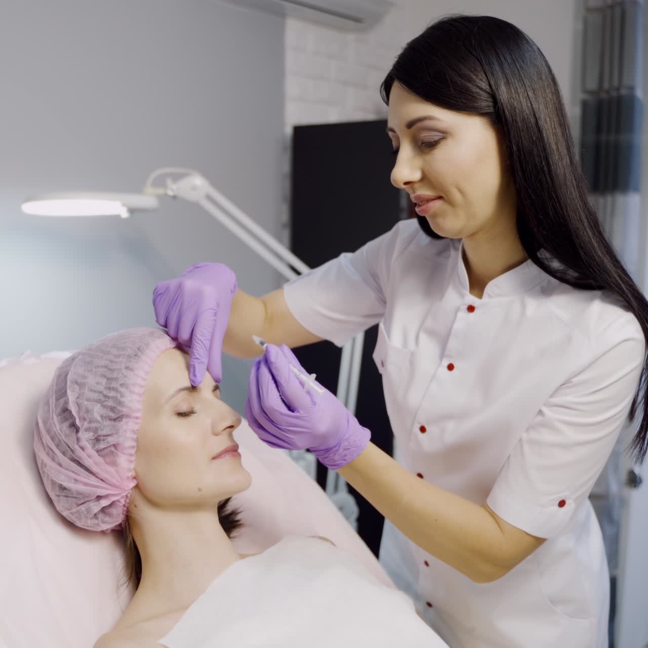 A pretty cosmetologist with a dark hair color makes Botox for a woman who lays on the couch in a beauty salon. Botulinum toxin