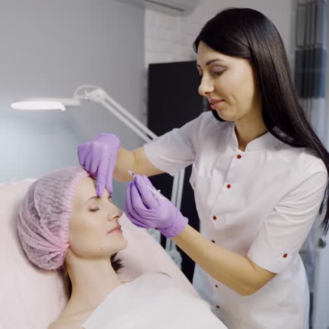 a pretty cosmetologist with a dark hair color makes botox for a woman who lays on the couch in a beauty salon. botulinum toxin