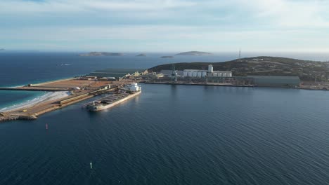 Toma-Aérea-Acercándose-Al-Puerto-Industrial-Con-Buque-Portacontenedores-Y-Fábrica-En-Segundo-Plano.